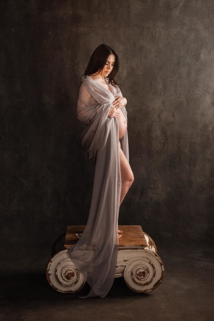 Fine art implied nude of pregnant woman on Restoration Hardware pedestal in front of gray handpainted backdrop with gray sheer fabric draped over her belly in an artistic manner