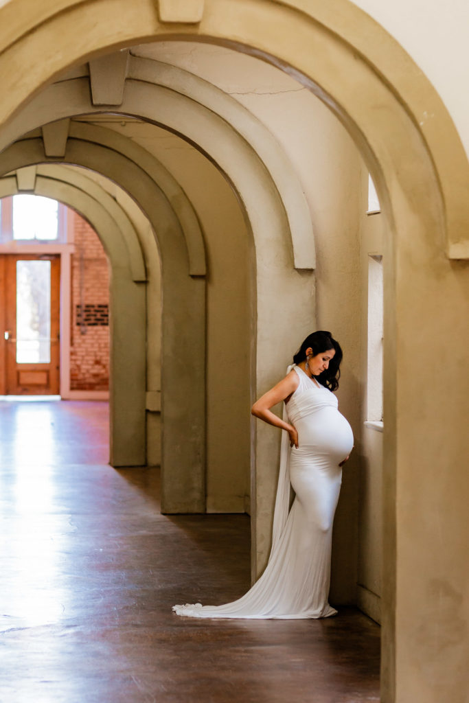 Brunette wearing white maternity gown at San Francisco Bay Area photo studio with arched hallway framing her as she gazes down at pregnant belly