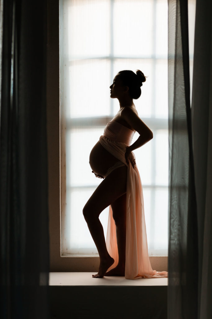 Silhouette photo of beautiful Asian pregnant woman wearing strapless blush pink sheer gown with belly exposed in studio for maternity photo session.