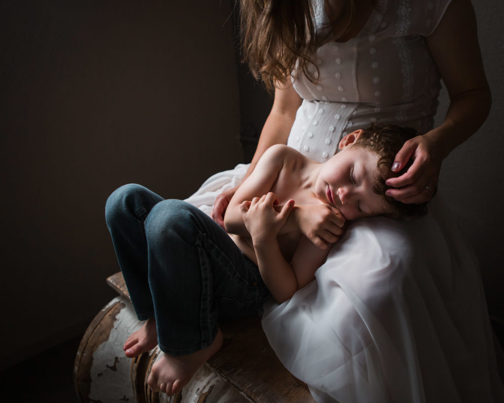 Fine art photo of pregnant mother holding toddler son in her lap. His eyes are closed and he is peaceful and serene