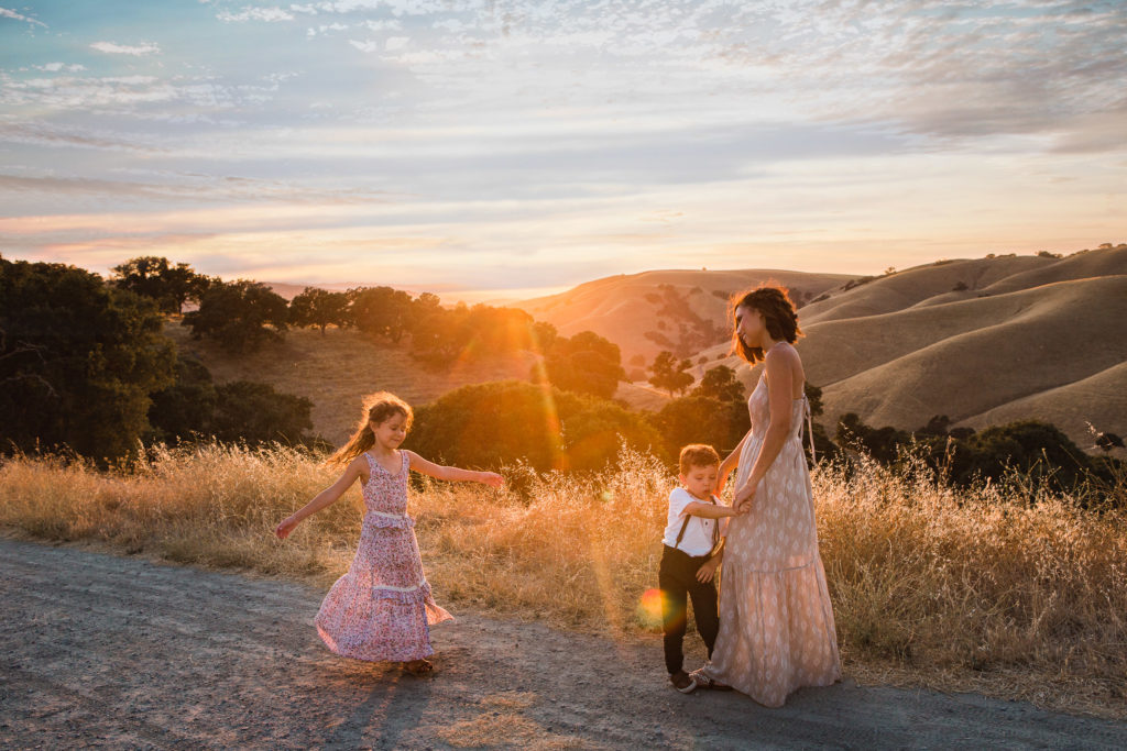 Little girl twirls during sunset photo session as little boy hangs on to mother who is smiling and looking at the children with golden northern california hills in the background