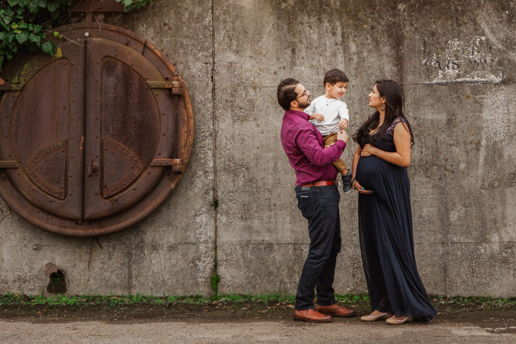 Family maternity photo session with casual dress. Parents holding toddler boy in front of concrete wall with greenery