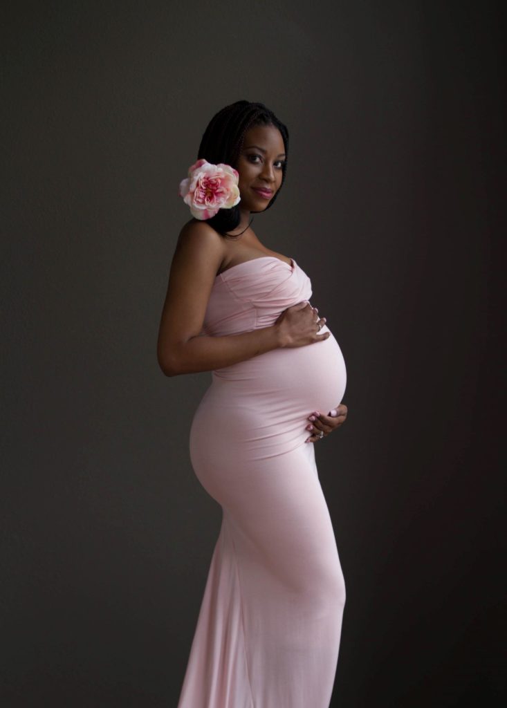Maternity portrait of African American woman wearing blush pink jersey fitted maternity gown with pink flower behind her ear.