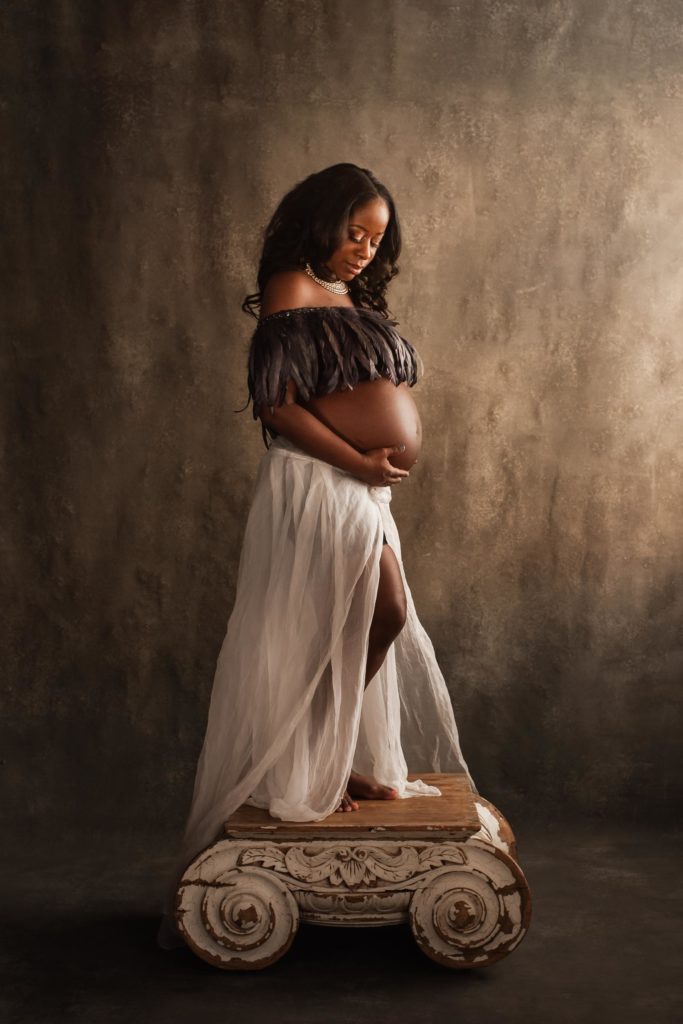 Pregnant African American woman standing on Restoration Hardware pedestal wearing feather top and tulle skirt in custom fine art maternity photo shoot.