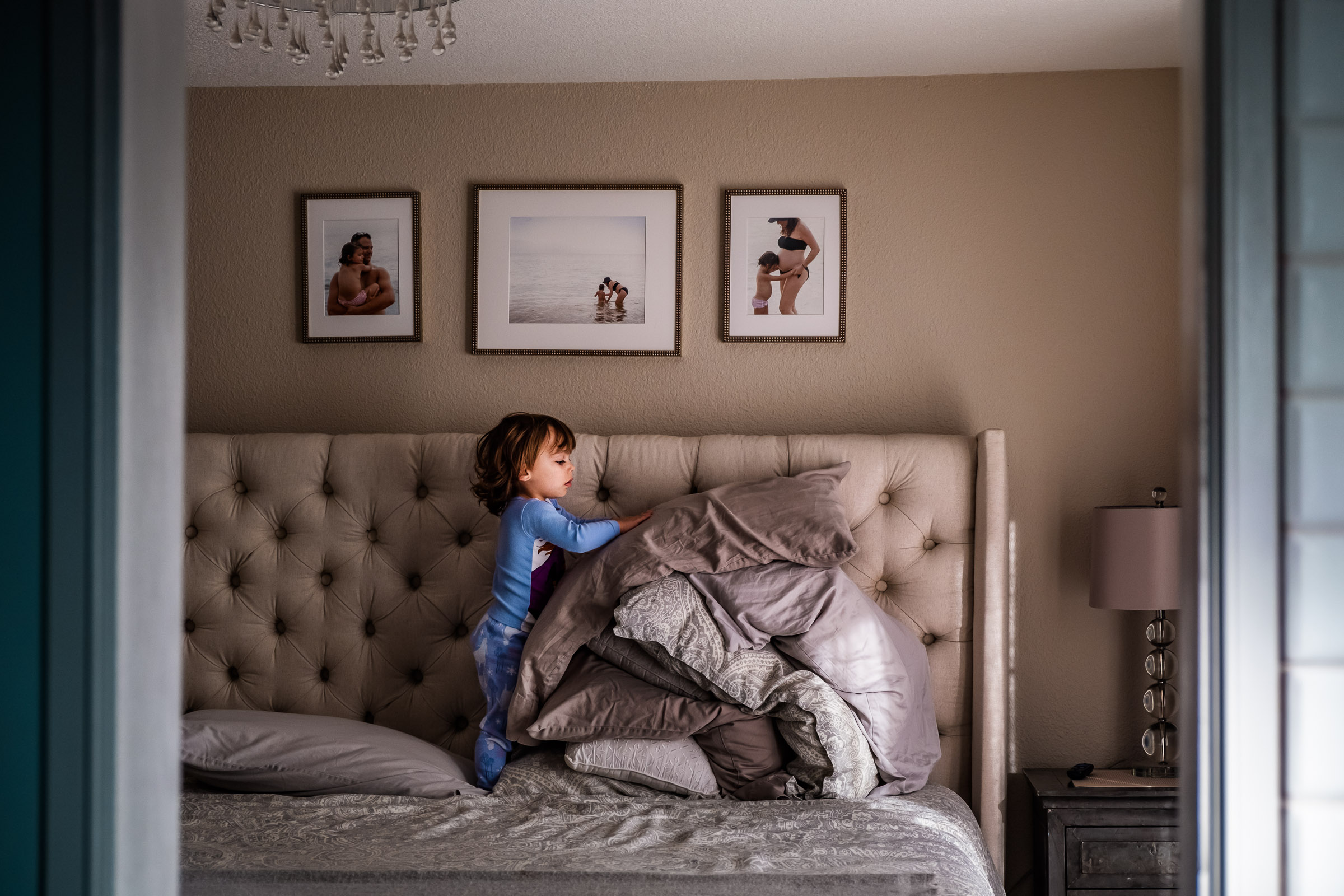 Toddler girl piles pillows on top of each other while she helps her mom make the bed.