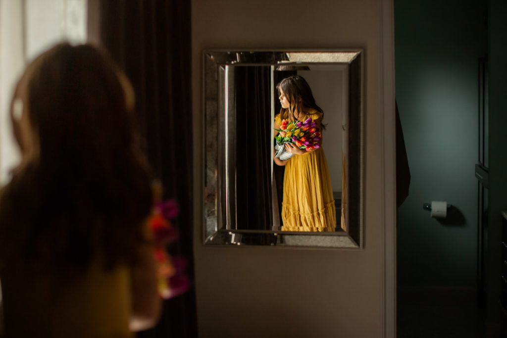 Artistic image of young girl in yellow dress, holding a bouquet of colorful tulips and looking out the window during COVID-19 pandemic with roll of toilet paper visible in the background.